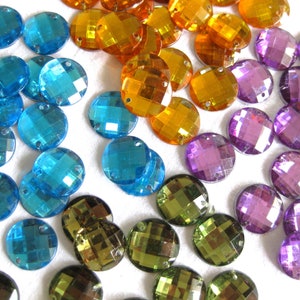 Könnte beinhalten: Sortiment runder, facettierter, irisierender Glas-Straßsteine in verschiedenen Farben, darunter Blau, Lila, Grün und Gold. Die Strasssteine sind auf einer weißen Oberfläche verstreut.