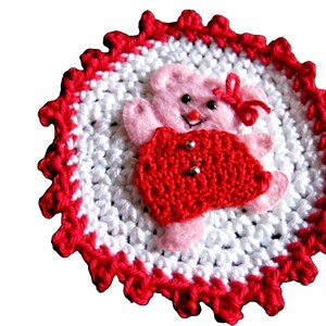 May include: A handmade crochet coaster featuring a pink teddy bear with a red dress and bow. The bear is centered on a white and red crocheted circle, ideal for a kitchen or dining room.