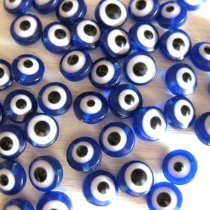 Peut inclure: Un ensemble de perles bleues et blanches en forme d'œil maléfique. Les perles sont rondes et ont un centre blanc avec une pupille noire. Les perles sont disposées de manière aléatoire sur une surface blanche.