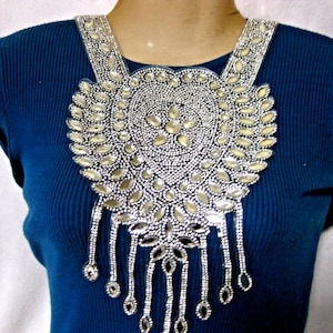 Op de afbeelding: Een zilveren kralenketting met een grote, traanvormige hanger. De hanger is gemaakt van gespiegeld glas en is omgeven door kleinere kralen. De ketting is op een blauwe achtergrond.