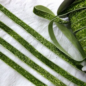 May include: Green glitter ribbon with a satin finish. The ribbon is rolled up and there are several strands of the ribbon laid out on a white surface.