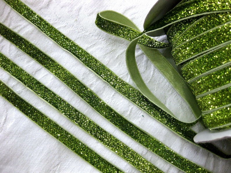 May include: Green glitter ribbon with a satin finish. The ribbon is rolled up and there are several strands of the ribbon laid out on a white surface.