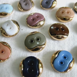 May include: A collection of round, two-hole buttons featuring a smooth, coloured centre inlay surrounded by a polished gold-tone metal rim. The buttons are available in various colours including white, black, blue, brown, pink, and grey.