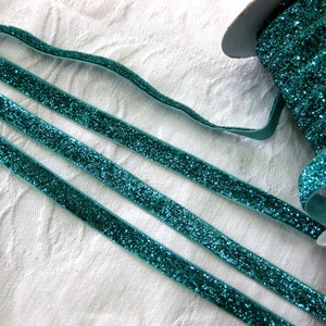 May include: Three strands of teal blue glitter ribbon on a white background. The ribbon is flat and has a velvet texture.