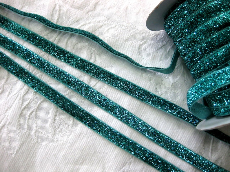 May include: Three strands of teal blue glitter ribbon on a white background. The ribbon is flat and has a velvet texture.