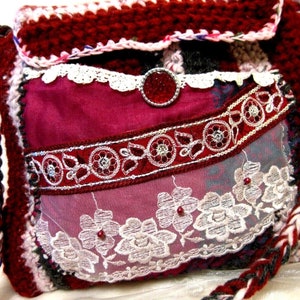 Może przedstawiać: Burgundowa torba na szydełku z białą koronkową lamówką i srebrno-czerwoną ozdobą z koralików. Torba ma pleciony pasek i czerwoną podszewkę.