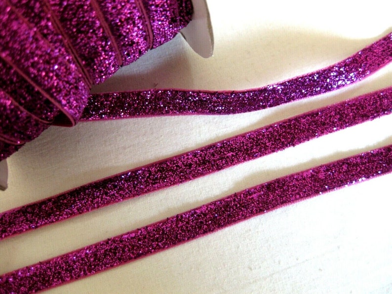 May include: Pink glitter ribbon with a velvet texture. The ribbon is rolled up and there are two strands of ribbon laid out flat.