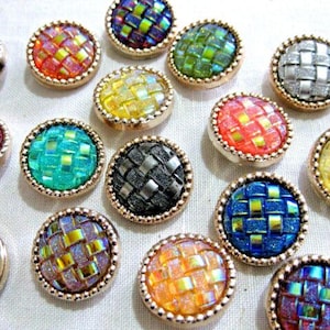 Op de afbeelding: Assortiment van kleurrijke, iriserende knopen met een geweven patroon. De knopen hebben een gouden metalen rand en zijn gerangschikt op een wit oppervlak.