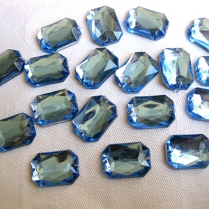 May include: A collection of rectangular, faceted, light blue glass rhinestones. Each stone has a flat back and is designed with multiple facets to catch and reflect light. The rhinestones are scattered on a white surface.