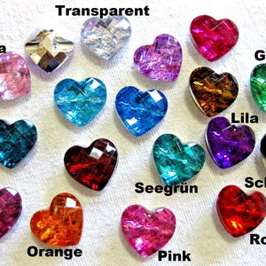 Könnte beinhalten: Eine Sammlung herzförmiger, facettierter Glasperlen in verschiedenen Farben, darunter Rosa, Orange, Rot, Grün und Blau. Das Wort "Transparent" steht über einer klaren herzförmigen Perle. Die Perlen sind auf einer weißen Oberfläche angeordnet.
