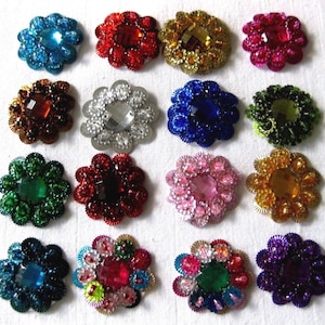 Puede incluir: Surtido de 16 adornos de colores en forma de flor con centros enjoyados. Cada adorno presenta un color y un diseño diferente, incluyendo plata, oro, rojo, azul, verde, rosa y morado.