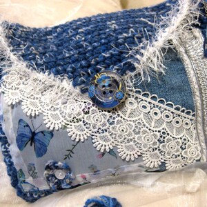 Può includere: Una borsa all'uncinetto blu e bianca con un bordo in denim e pizzo. La borsa ha un bordo in piume bianche e un bottone blu e bianco con un motivo floreale.