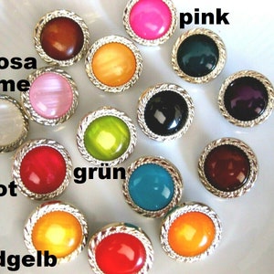Peut inclure: Assortiment de 14 boutons ronds avec une bordure argentée et de différentes couleurs, notamment rose, rouge, vert, or, marron, noir et blanc. Les boutons sont disposés en cercle sur une surface blanche.