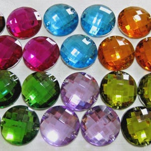 Peut inclure: Collection de vingt-et-un bijoux ronds et facettés de différentes couleurs. Les couleurs comprennent le marron, le fuchsia, le bleu, l'orange, le vert, le violet et le jaune-vert. Chaque bijou a un dos plat et un trou pour la fixation.