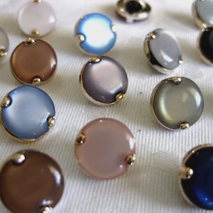 May include: A collection of round buttons in various colors, including blue, gray, brown, and pink. Each button has a gold-colored rim and two small gold accents. The buttons are arranged on a white surface.