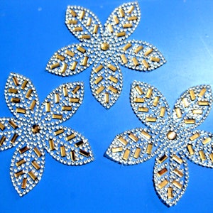 May include: Three decorative flower appliques. Each flower has five petals, with a silver outline and filled with small, clear rhinestones. Gold rectangular accents are interspersed. The flowers are set against a bright blue background.