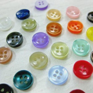 Può includere: Una collezione di bottoni rotondi e colorati, ciascuno con quattro fori. I bottoni sono in varie tonalità, tra cui blu, verde, giallo, arancione e viola. Sono sparsi su una superficie bianca, mostrando i loro diversi colori e design.