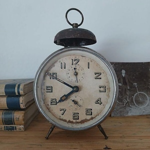 Könnte beinhalten: Ein antiker Wecker mit cremefarbenem Zifferblatt, schwarzen Ziffern und schwarzen Zeigern. Die Uhr hat einen Metallrahmen, eine Glocke oben und drei kleine Beine. Die Uhr steht auf einer Holzoberfläche.