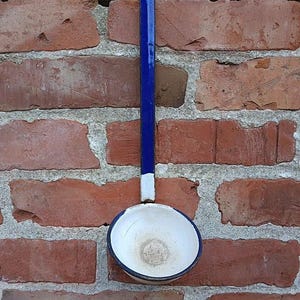 Könnte beinhalten: Eine blaue und weiße Emaille-Schöpfkelle hängt an einer roten Backsteinmauer. Die Schöpfkelle hat einen langen, geraden blauen Griff und eine runde, weiße Schüssel mit blauem Rand. Die Backsteinmauer besteht aus verwitterten roten Ziegeln.