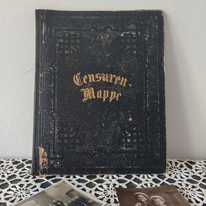 Könnte beinhalten: Ein schwarzes, ledergebundenes Buch mit goldenen Lettern, auf dem "Censuren-Mappr" steht. Das Buch liegt auf einer weißen Spitzentischdecke mit zwei Schwarzweißfotos.