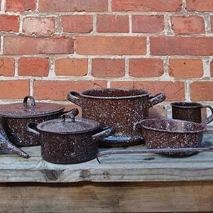 Könnte beinhalten: Eine Sammlung von Vintage-Küchenartikeln aus braun gesprenkeltem Emaille, darunter Töpfe, ein Trichter, eine Schöpfkelle und eine Tasse, angeordnet auf einer Holzoberfläche vor einer Backsteinmauer. Das Kochgeschirr hat ein gesprenkeltes Muster.