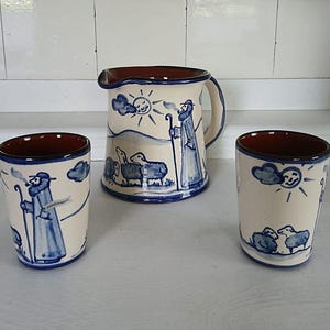 Könnte beinhalten: Ein Keramikkrug und zwei passende Becher. Die cremefarbenen Stücke zeigen eine blaue pastorale Szene mit einem Hirten, Schafen, Sonne und Wolken. Die Ränder und das Innere sind braun. Der Krug hat einen Henkel und einen Ausguss.