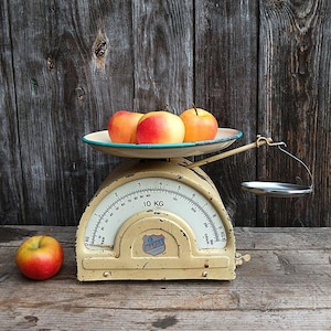 Könnte beinhalten: Eine antike cremefarbene Waage mit weißem Zifferblatt und einem blau umrandeten Tablett mit drei rot-gelben Äpfeln. Ein einzelner Apfel liegt auf der Holzoberfläche. Die Waage zeigt bis zu 10 kg an.