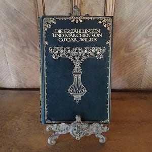 Könnte beinhalten: Ein gebundenes Buch mit dem Titel "Die Erzählungen und Märchen von Oscar Wilde" in deutscher Sprache, auf einem dekorativen Metallständer. Das Buch hat einen dunkelblauen Einband mit goldenen Verzierungen und Schrift. Der Ständer ist hellgrau.