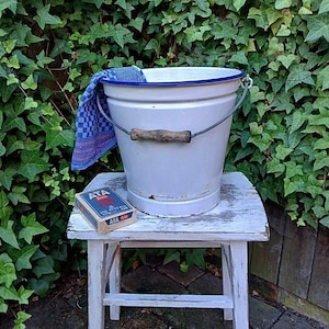 Könnte beinhalten: Ein weißer Emaille-Eimer mit blauem Rand und Holzgriff steht auf einem kleinen, weißen, abgenutzten Hocker. Ein blau-weiß kariertes Handtuch hängt über dem Eimerrand. Eine Schachtel "ATA fein" liegt auf dem Hocker.