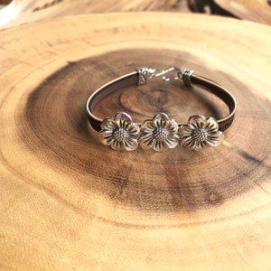 Op de afbeelding: Een zilveren armband met drie bloembedels. De armband heeft een bruine leren band en een zilveren sluiting. De bloembedels zijn gedetailleerd met ingewikkelde bloemblaadjes, waardoor een delicaat en vrouwelijk accessoire ontstaat.