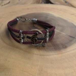 Könnte beinhalten: Ein Armband aus burgunderfarbenem und schwarzem Leder mit einem Treble-Clef-Anhänger und silbernen und goldenen Draht-Akzenten.