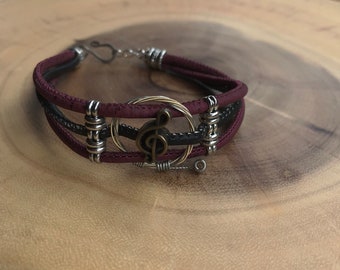 May include: A burgundy and black leather bracelet with a treble clef charm and silver and gold wire accents.