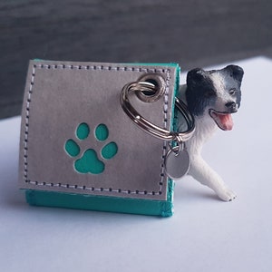 Könnte beinhalten: Ein grauer und türkisfarbener Leder-Schlüsselanhänger mit einem Pfotenabdruck-Ausschnitt und einem silbernen Schlüsselring. Eine kleine Hundefigur ist teilweise sichtbar, die hinter dem Schlüsselanhänger hervorlugt. Ein Accessoire zum Thema Haustiere.