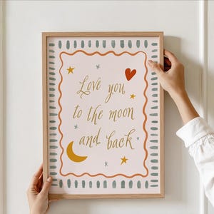 以下が含まれることがあります： 「Love you to the moon and back」の文字が金色のフレーム入りアートプリント。ピンク色の背景に赤いハート、黄色の星、三日月が描かれています。フレームは明るい茶色です。
