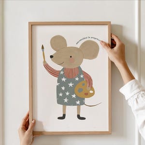Może przedstawiać: Oprawiony wydruk przedstawiający mysz z kreskówki, artystę trzymającą pędzel i paletę. Mysz nosi fartuch we wzór gwiazd i koszulę w paski. Tekst "masterpiece in progress" jest wygięty nad myszą.