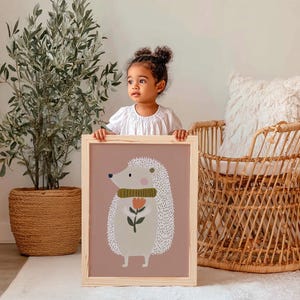 May include: A framed art print featuring a cartoon hedgehog with a flower. The hedgehog has white quills, a green scarf, and a pink nose. The background is a muted rose color, and the frame is natural wood.