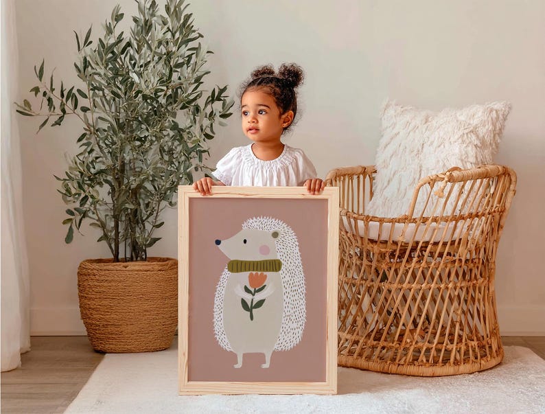 May include: A framed art print featuring a cartoon hedgehog with a flower. The hedgehog has white quills, a green scarf, and a pink nose. The background is a muted rose color, and the frame is natural wood.