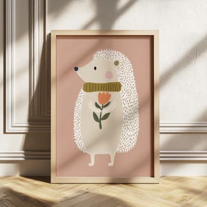 May include: A framed illustration of a hedgehog holding a flower. The hedgehog has white quills, a green scarf, and a pink cheek. The background is a solid dusty rose color, and the frame is light wood.