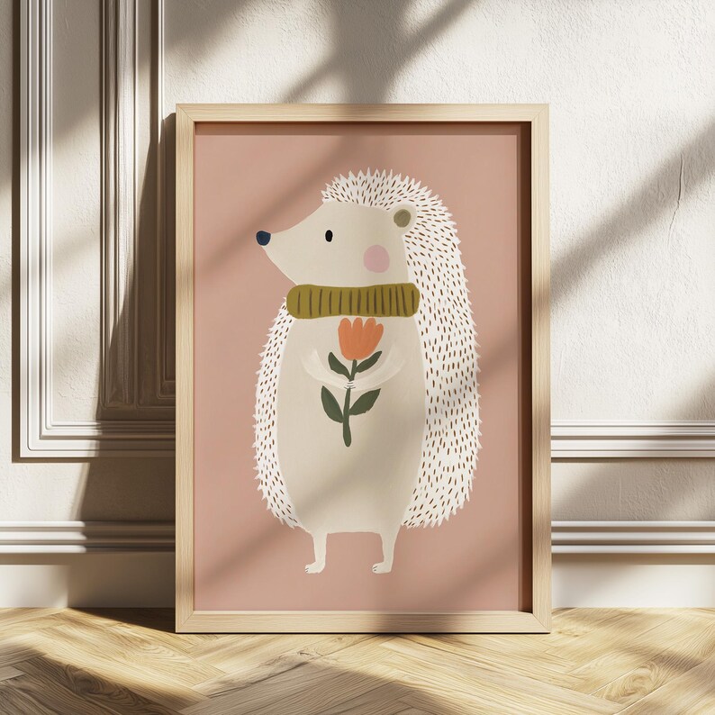 May include: A framed illustration of a hedgehog holding a flower. The hedgehog has white quills, a green scarf, and a pink cheek. The background is a solid dusty rose color, and the frame is light wood.