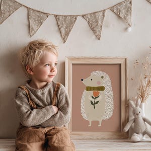 May include: A framed print featuring a whimsical hedgehog illustration with a green scarf and a flower. The artwork is in a light wooden frame, set against a muted pink background. A child is sitting next to the framed print.