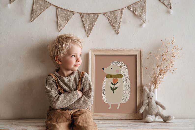 May include: A framed print featuring a whimsical hedgehog illustration with a green scarf and a flower. The artwork is in a light wooden frame, set against a muted pink background. A child is sitting next to the framed print.