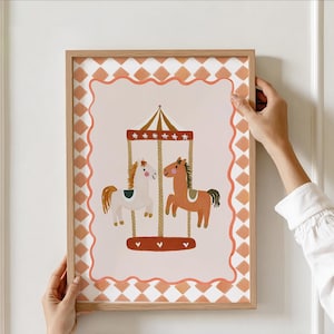 May include: A framed art print depicting a carousel scene with two horses. The horses are cream and brown, with a carousel top and base in shades of red and brown. The print has a peach-coloured background and a decorative border.
