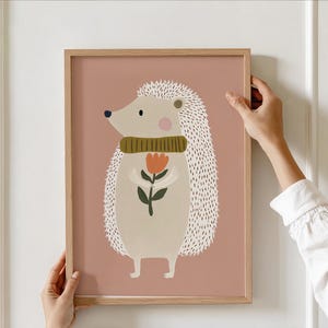 May include: A framed art print featuring a whimsical illustration of a hedgehog. The hedgehog is depicted with a cream-colored body, a green scarf, and is holding a flower. The background is a muted rose color, and the frame is a light wood.