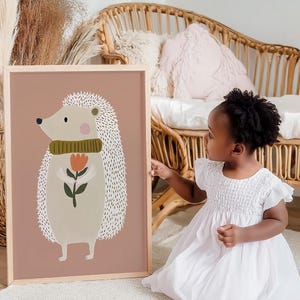 May include: A framed art print featuring a whimsical illustration of a hedgehog holding a flower. The hedgehog has a light beige body, a green scarf, and a pink cheek. The background is a muted rose color.