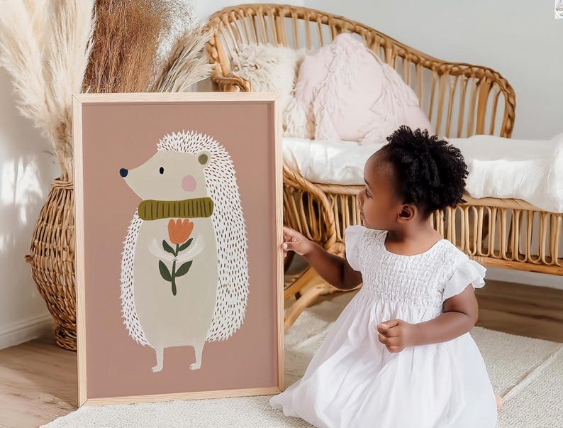 May include: A framed art print featuring a whimsical illustration of a hedgehog holding a flower. The hedgehog has a light beige body, a green scarf, and a pink cheek. The background is a muted rose color.
