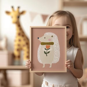 May include: A framed art print featuring a whimsical illustration of a hedgehog holding a flower. The hedgehog has a cream-colored body, a green scarf, and a light brown frame. The background is a soft pink.