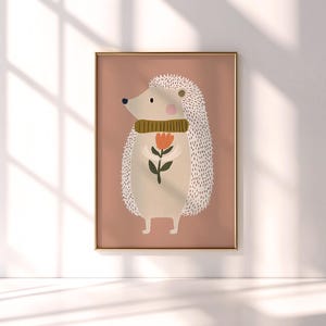 May include: A framed art print featuring a whimsical illustration of a hedgehog. The hedgehog is depicted with a beige body, white quills, and a green scarf, holding a red and orange flower. The background is a muted pink.