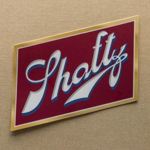 May include: A rectangular sticker with the word "Shatty" in a stylized font. The text is white with a blue outline, set against a maroon background. The sticker has a gold border and is set against a tan background.