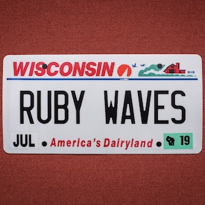 May include: A white Wisconsin license plate with the text "RUBY WAVES" in black lettering. The plate also features the text "JUL - America's Dairyland - 19" in black lettering. The top of the plate has the text "WISCONSIN" in red lettering with a red circle and a red barn graphic.