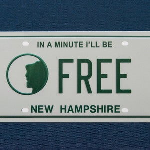 May include: A white metal license plate with green text and a green silhouette of a person's head in a circle. The text reads "In a minute I'll be FREE New Hampshire".